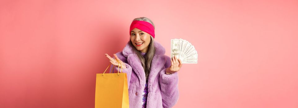 Rich and stylish grandmother going on shopping with dollars, holding paper bag and smiling carefree, standing over pink backgrund.