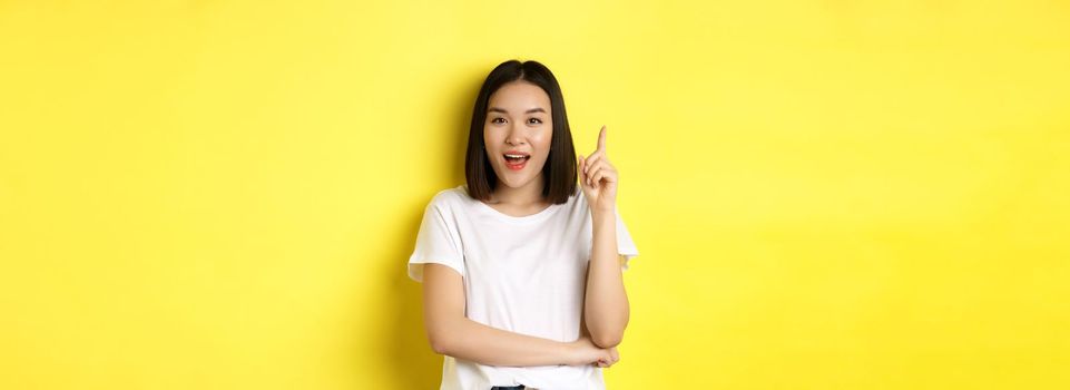 Beauty and fashion concept. Excited asian girl raising finger in eureka gesture, pitching an idea and smiling, standing over yellow background.