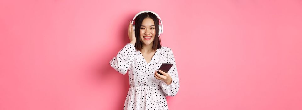 Beautiful asian woman listening music in headphones, using smartphone streaming app and smiling, standing over pink background.