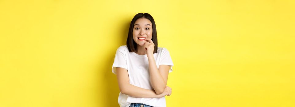 Portrait of cheerful asian girl wants something, looking with desire and temptation at camera, smiling at camera, standing ovr yellow background.