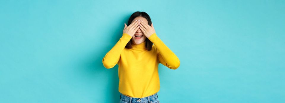 Cheerful asian girl in yellow pullover waiting for surprise, playing hide n seek and smiling, expecting gift with eyes closed, standing over blue background.