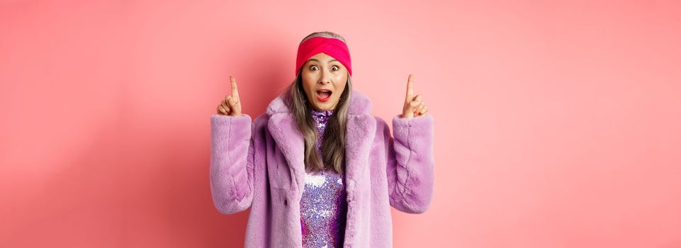 Happy elderly asian lady pointing fingers up, looking amazed at camera, showing cool promo offer, standing against pink background in purple fake fur coat.