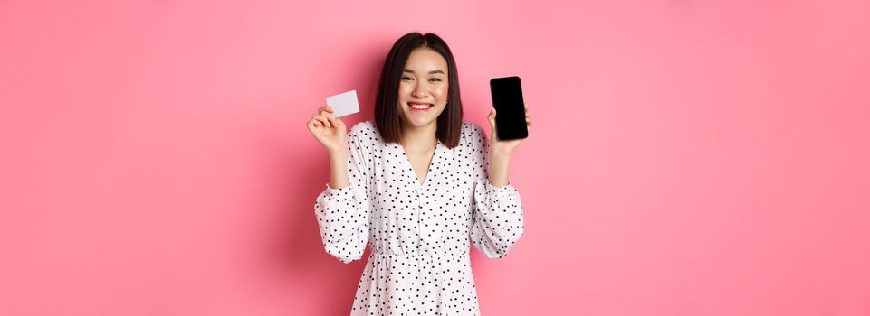 Joyful korean woman showing smartphone screen and credit card, paying for internet order, demonstrating online shopping app, standing over pink background.