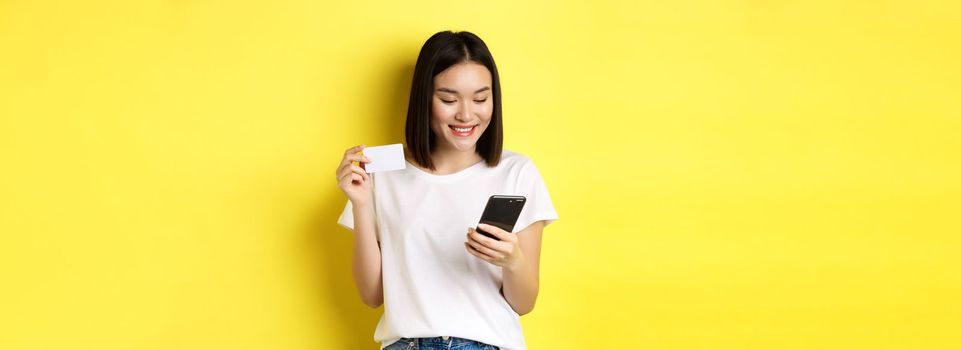 E-commerce and online shopping concept. Beautiful asian woman paying in internet, looking at smartphone screen and holding plastic credit card, yellow background.