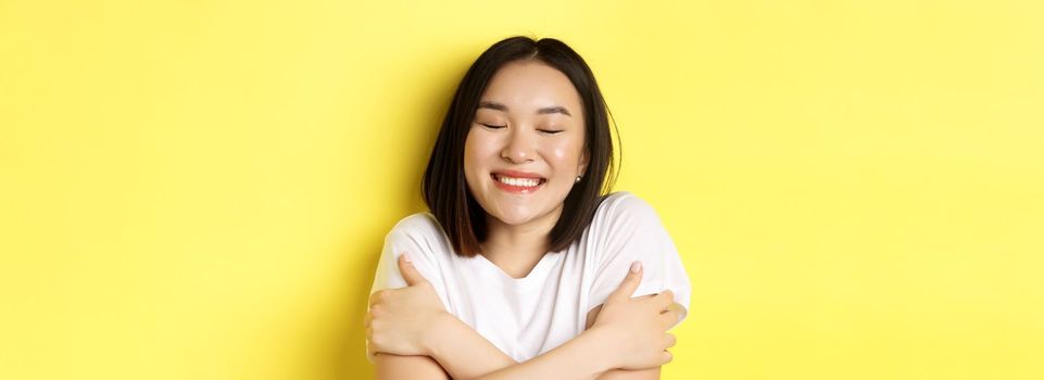 Close up of romantic asian girl hugging herself and dreaming, close eyes and smile while imaging something tender, standing over yellow background.