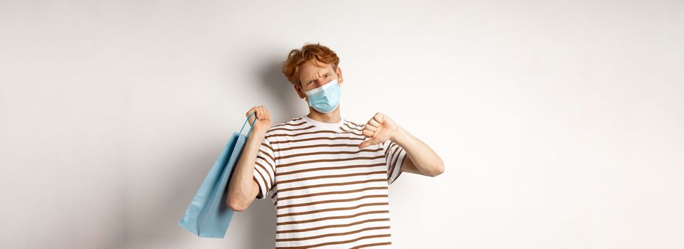 Concept of social distancing and shopping. Disappointed young man with red hair, wearing face mask, holding shopping bag and showing thumb-down from dislike, disapprove store.