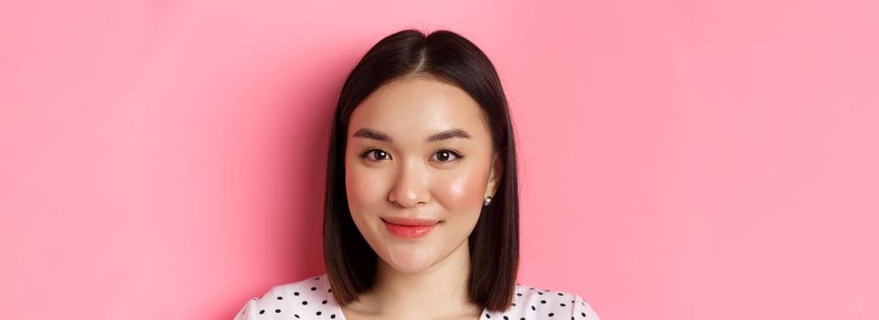 Beauty and lifestyle concept. Headshot of beautiful asian woman smiling, looking at camera happy and romantic, standing against pink background.