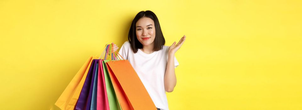 Silly asian girl showing shopping bags and shrugging, shop in stores with discounts, standing over yellow background.