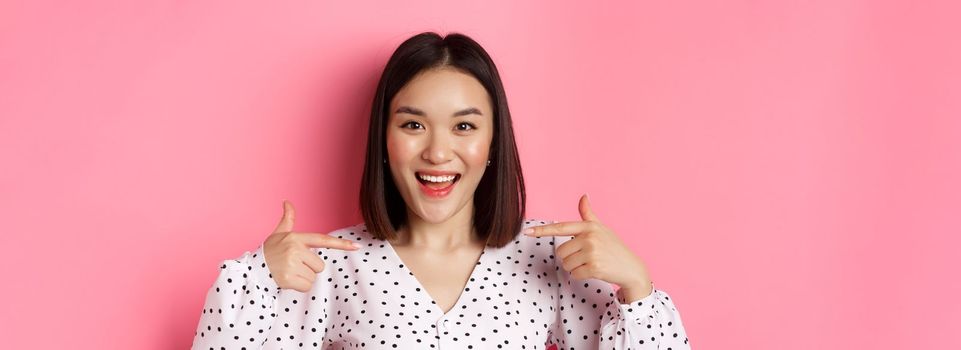 Close-up of happy and confident asian woman pointing at herself, smiling self-assured, standing over pink background.