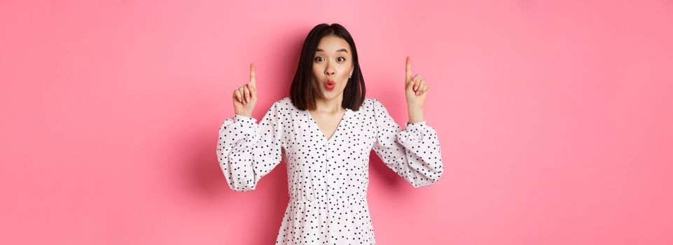 Pretty asian female model in dress pointing fingers up, showing copy space and staring at camera amazed, telling big news, standing over pink background.