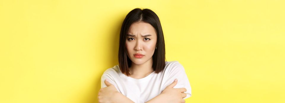 Sad timid girl comforting herself, hugging body and frowning upset, standing again yellow background.