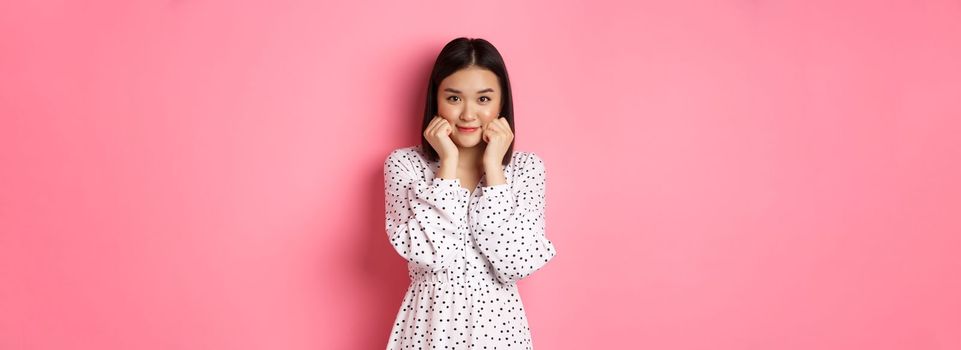 Cute and shy asian girl blushing, touching cheeks and looking at camera silly, standing against pink background.