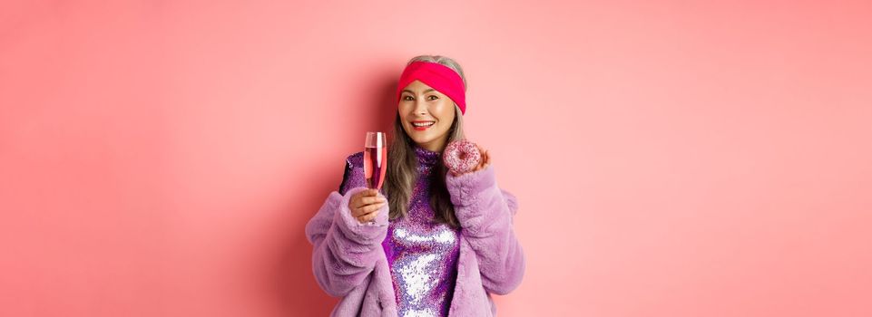 Elegant and stylish asian woman in purple faux fur coat eating donuts and drinking champagne, having fun at party, standing over pink background.