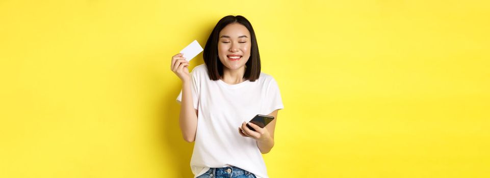 E-commerce and online shopping concept. Happy asian woman looking excited, buying something in internet, holding smartphone and showing plastic credit card, yellow background.