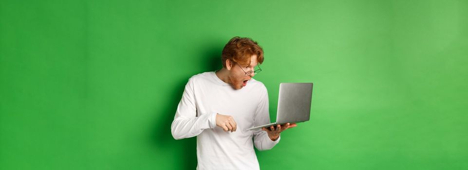 Image of young redhead man looking surprised at laptop screen, drop jaw and staring at display amazed, green background.