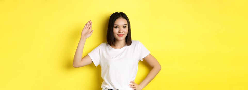 Friendly asian woman in white t-shirt waving hand and saying hello, greeting you, standing over yellow background.