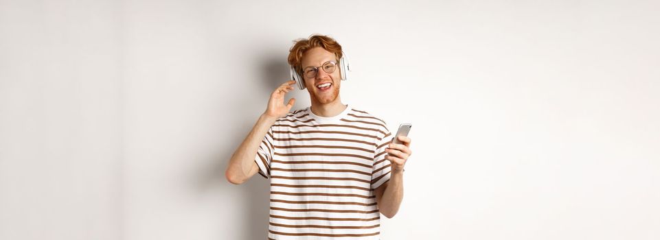 Technology concept. Young man with red hair and beard listening music in headphones and using smartphone, smiling at camera, white background.