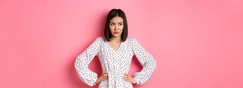 Sad and concerned asian woman having problem, holding hands on waist and looking left with upset face, standing in dress against pink background.