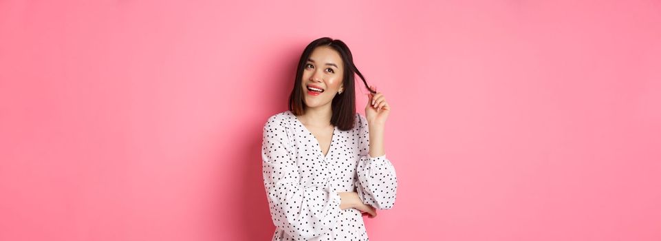 Dreamy cute asian girl thinking, imaging shopping and smiling, looking upper left corner with thoughtful smile, standing in dress over pink background.