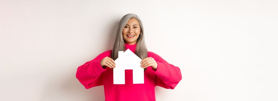 Real estate. Beautiful asian grandmother showing paper house and smiling happy, standing in pink sweater over white background.