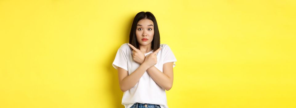 Indecisive asian girl need help with choice, pointing fingers sideways and looking confused, standing over yellow background.