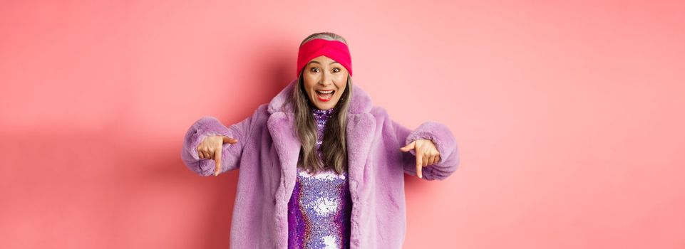 Fashion and shopping concept. Happy korean senior woman pointing fingers down and showing awesome promo deal, wearing trendy purple fur coat and headband, pink background.