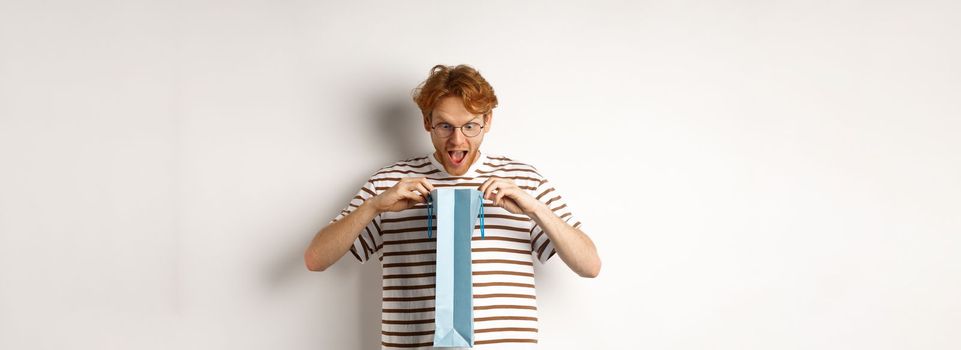 Surprised boyfriend open shopping bag with valentines day gift, staring inside with amazed face, standing over white background.