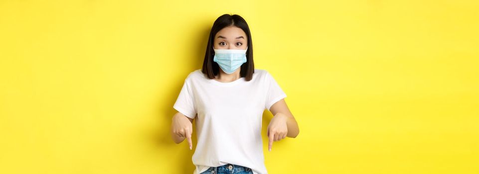 Covid-19, pandemic and social distancing concept. Amazed asian woman in medical mask, showing advertisement, pointing right and smiling, yellow background.