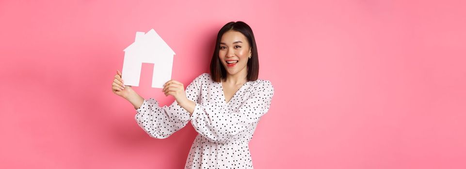 Real estate. Beautiful asian woman demonstrating paper house model, looking at camera confident, advertising home for sale, standing over pink background.