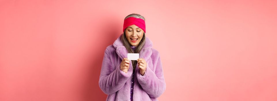 Shopping and fashion concept. Beautiful and stylish asian woman looking at plastic credit card, smiling pleased, standing over pink background.