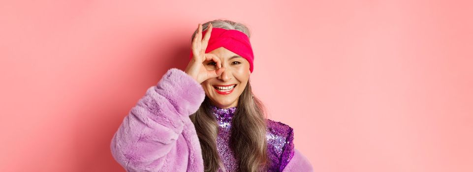 Fashion. Close-up of fashionable asian senior woman smiling, showing OK sign over eye and looking happy at camera, standing over pink background.