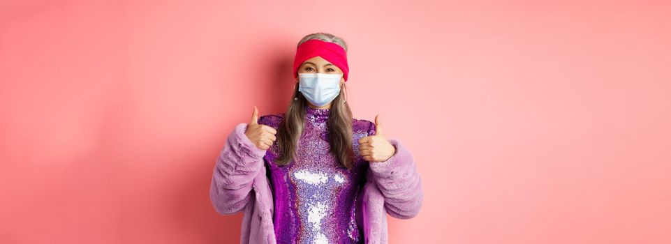 Coronavirus and shopping concept. Stylish asian grandmother in respirator and glittering purple dress and faux fur coat, showing thumbs-up, recommend shop in face masks.