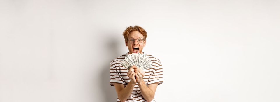 Lucky young man with red hair showing dollars, winning money and screaming of happiness, holding prize cash, standing over white background.