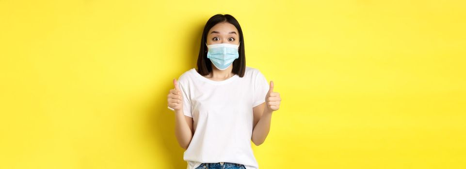 Covid-19, quarantine and social distancing concept. Cheerful asian woman in medical mask and white t-shirt showing thumbs up in approval, praise good deal, yellow background.