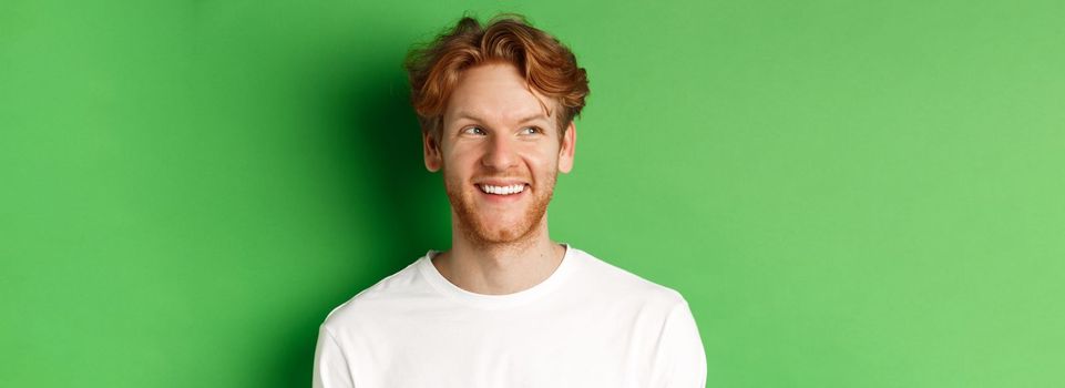 Headshot of happy redhead man with beard wearing white long sleeve, looking left at copy space and smiling, standing over green background.
