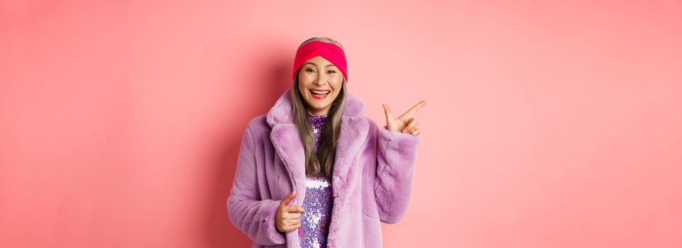 Fashion and shopping concept. Happy asian senior woman checking out trendy promotion, pointing finger left at logo and smiling, wearing winter fake fur coat and headband.