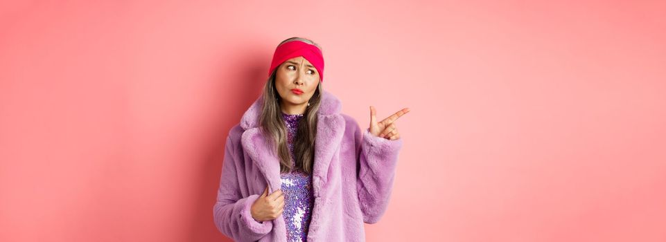 Fashion and shopping concept. Skeptical middle-aged asian woman in purple fur coat looking and pointing finger left with displeased, sad expression, standing over pink background.