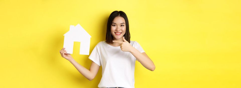Real estate and insurance concept. Cheerful asian woman smiling, pointing at paper house cutout, recommend agency logo, standing over yellow background.