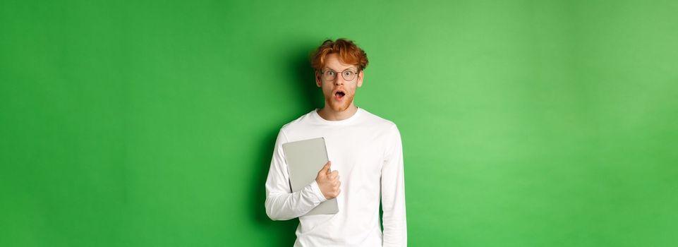 Surprised young man holding laptop and staring at camera, wearing glasses and white t-shirt, standing over green background.