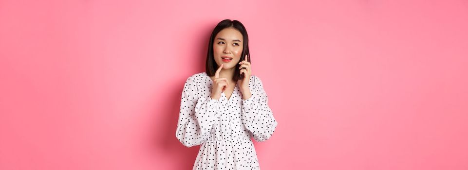 Thoughtful asian woman calling on mobile phone to make order, thinking and looking aside, standing against pink background.