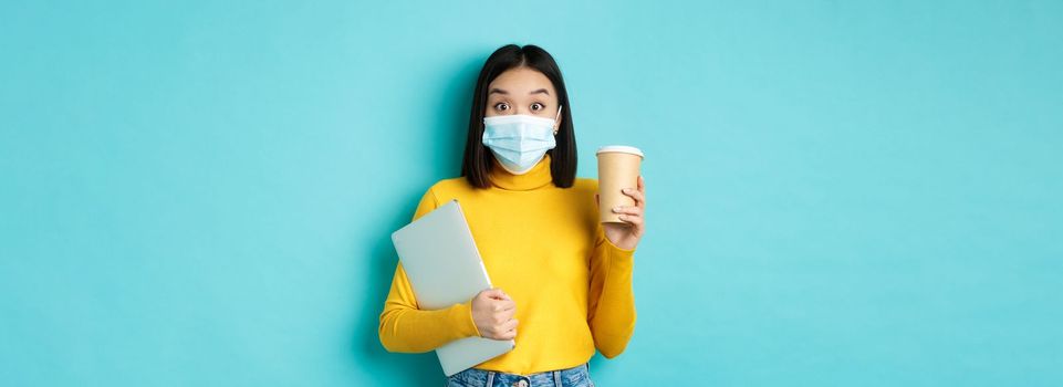covid-19, health care and quarantine concept. Asian girl student in medical mask standing with laptop and coffee from cafe, standing over blue background.