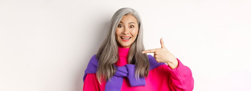 Close up of beautiful and stylish asian senior woman smiling, pointing at herself, showing logo, standing over white background.