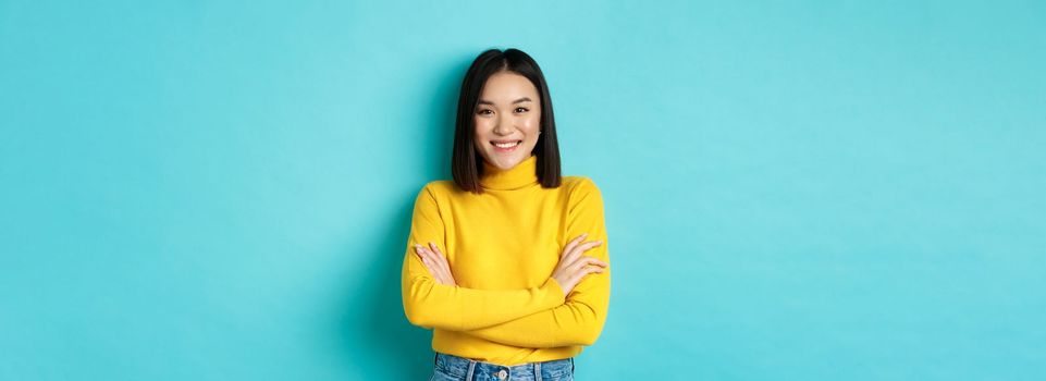 Confident and stylish asian woman cross arms on chest and smiling, standing over blue background.