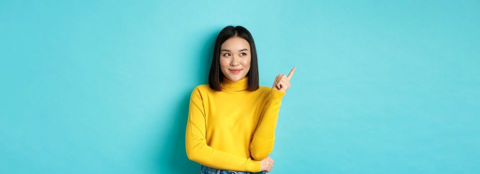 Shopping concept. Stylish asian female model in yellow sweater, smiling and pointing finger left, showing advertisement with satisfied face, standing over blue background.