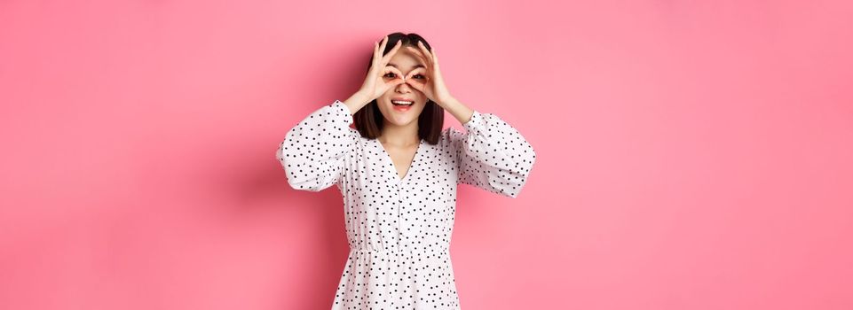 Carefree young asian woman looking through hand binoculars at camera, staring at discounts, standing over pink background.