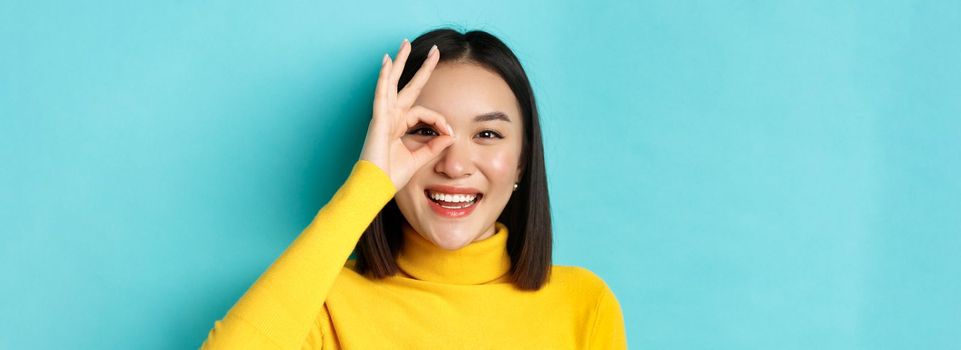 Beauty and makeup concept. Close up of carefree asian girl showing OK sign on eye and smiling, looking happy at camera, standing over blue background.