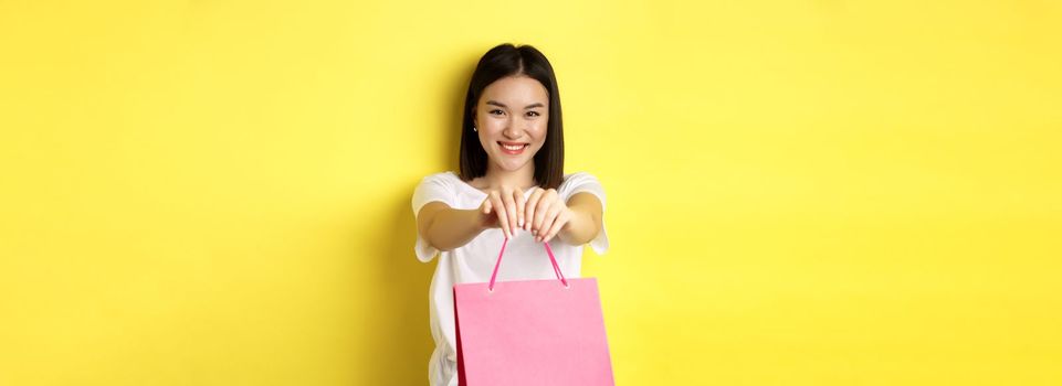 Cute asian girl giving you gift, stretch out hand with pink shopping bag and smiling, congratulating with holiday, standing over yellow background.