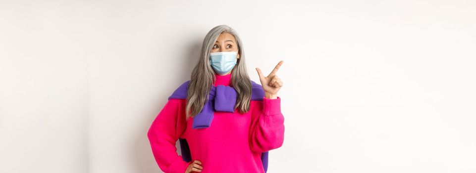 Covid-19, quarantine and lockdown concept. Amazed asian senior woman in face mask looking impressed, showing logo on upper left corner, pointing at banner, white background.