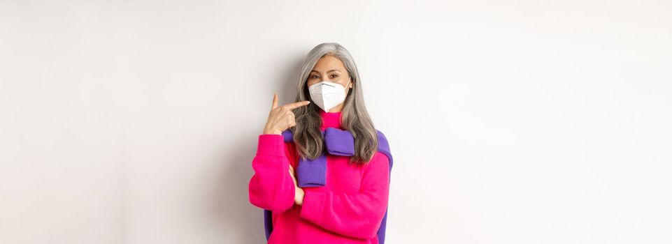 Covid, pandemic and social distancing concept. Fashionable asian senior woman wearing respirator, pointing at face mask and smiling, standing over white background.