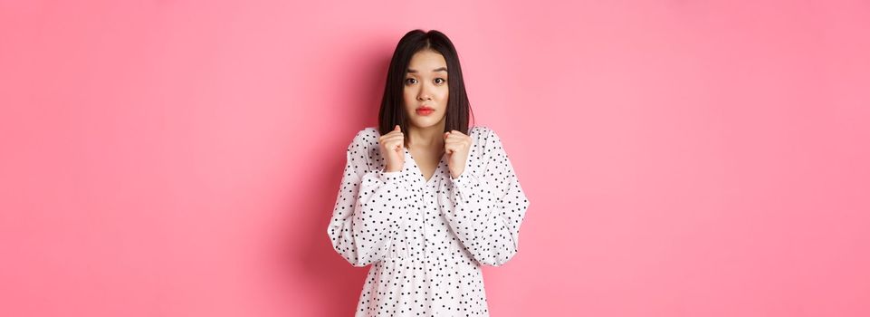 Scared cute asian girl in dress looking worried, feeling frightened and staring at camera nervously, standing over pink background.